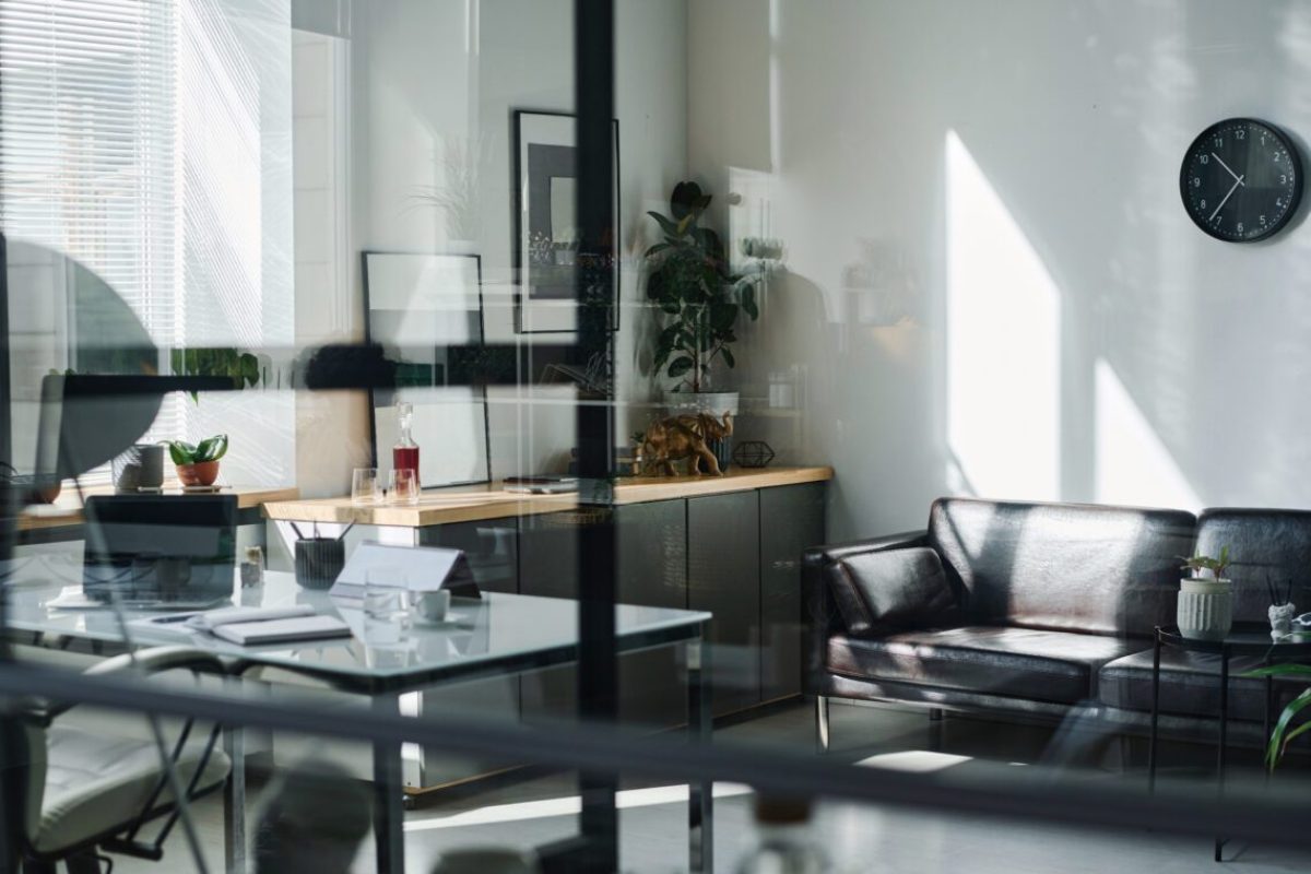 Focus on wall with clock hanging over comfortable black leather couch standing in the corner of spacious modern office with green plants