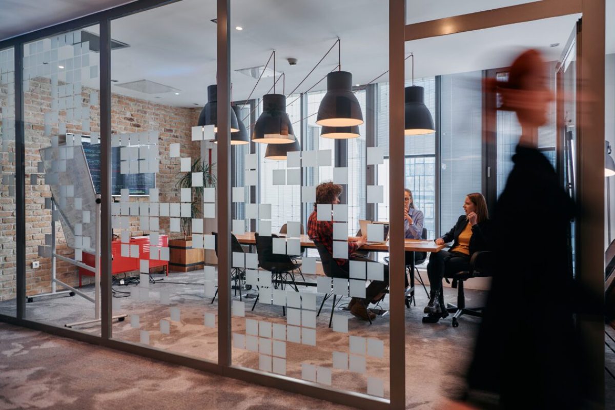 In the dynamic hustle and bustle of a business environment, a group of young business professionals walk down the corridor next to their office, while their colleagues collaborate inside, symbolizing teamwork and productivity.