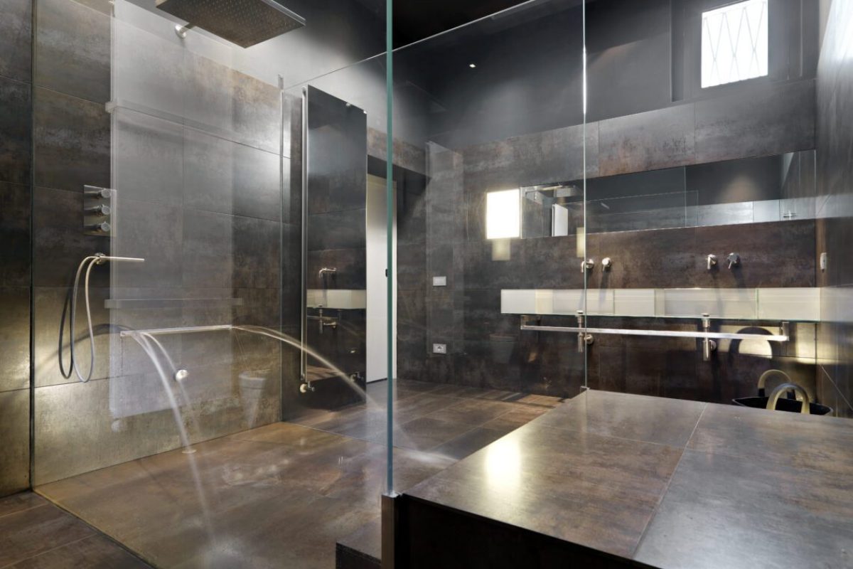 view a large masonry shower cubicle and masonry bathtub  with glass partition and dark tiled overlooking on the backgroound the glass washbasin