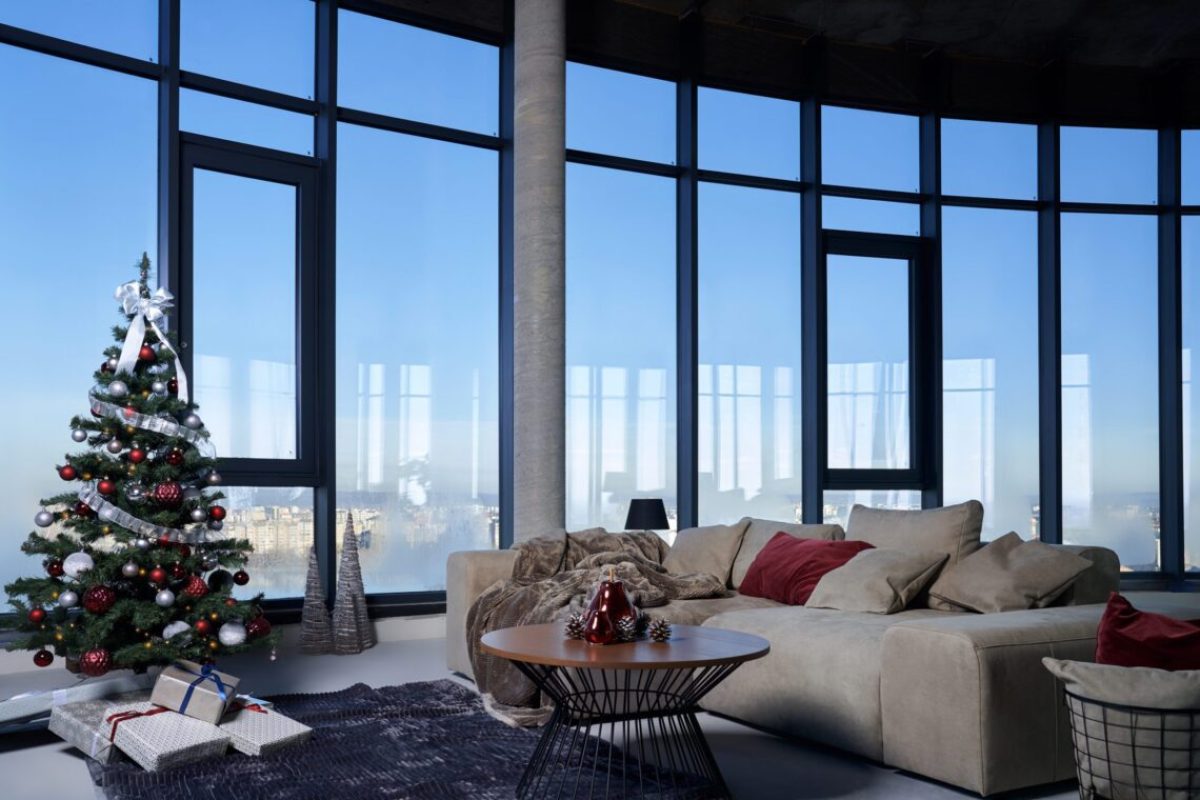 Front view of cozy grey sofa with pillows near beautiful Christmas tree and large panoramic window with incredible view. Concept of interior modern, spacious room.