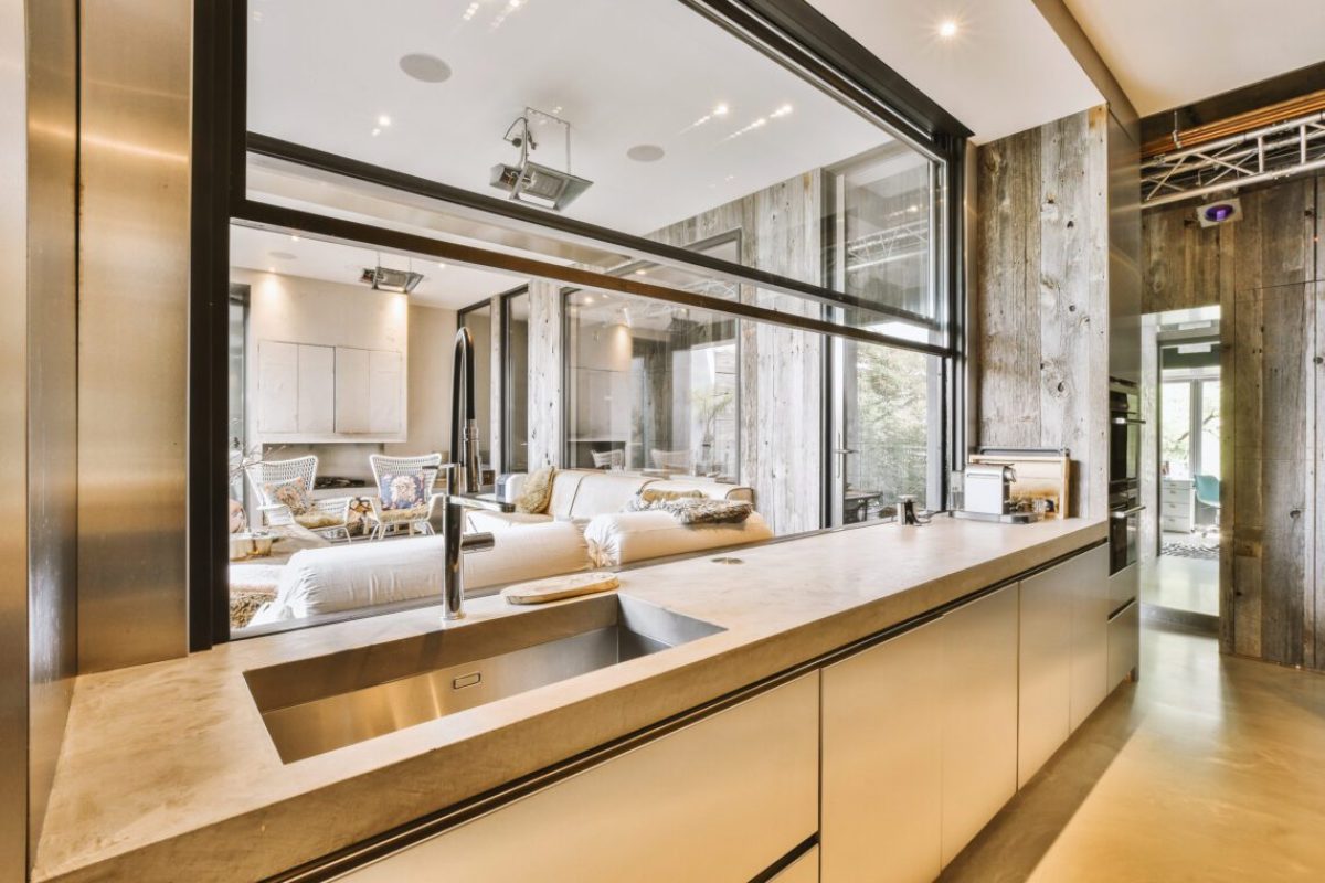 Large corner sofa with soft cushions located under elegant chandelier near electric fireplace in light living room near kitchen