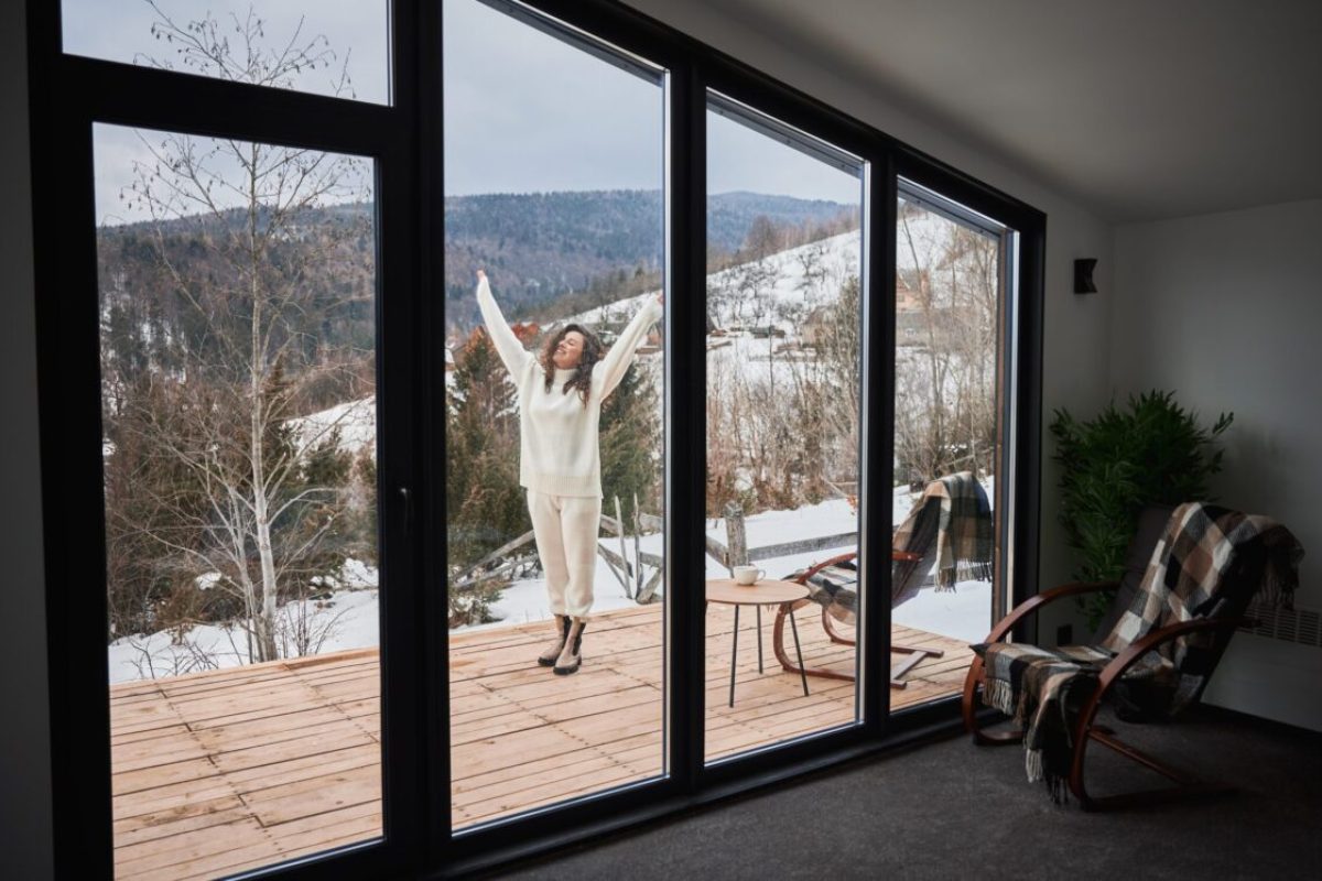Curly young woman resting on terrace of modern barn house in the mountains. View from inside cottage. Happy female tourist enjoying winter holiday.